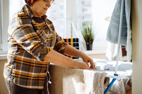 Side view of senior woman wearing checkered shirt, brown trousers, standing at rack dryer with different clothes. Woman putting hands on clothes-horse on sunny day. Household chores, housekeeping.