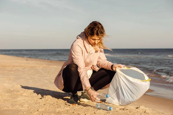 Woman activist and assistant cleaning sandy beach from underwater trash, sea or ocean with plastic waste. Collect bottles to bag. Ecological pollution. Sustainable development. Environmental campaign