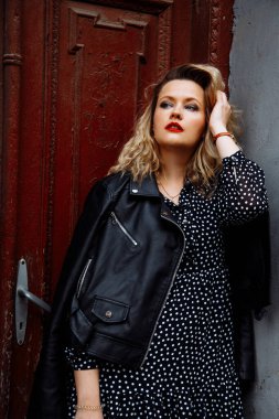 Modern gülümseyen stil sarışın kadının dikey görüntüsü. Makyajlı, siyah deri ceketli ve kahverengi ev kapısına dayanmış bir kadın. Şehir tarzı. Kadınsı cazibe, yeni bir hayat yolculuğuna başla.