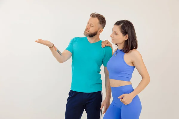 Young athletic woman and man in blue sportswear standing and pointing by hand, white background. Keeping fit by fitness workout in gym, pumping up body. Family sport and healthy lifestyle concept.