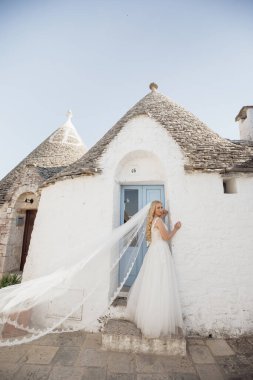 Beyaz gelinlik ve duvak takmış, gülümseyen ve İtalya 'da taş şehir evinin yanında duran genç sarışın gelin portresi. Güzel ve romantik bir düğün, sokakta yürümek..