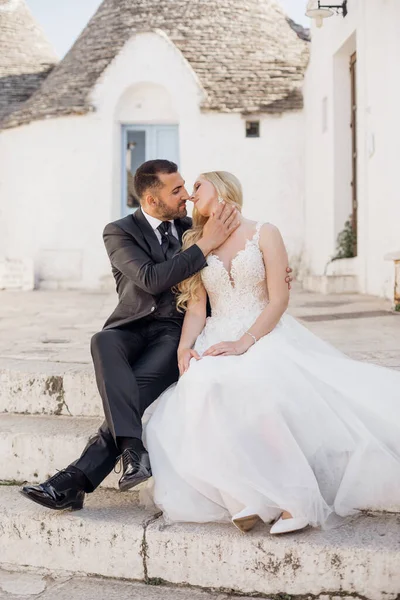 白いドレスを着た若いブロンドの花嫁とスーツを着たブルネットの新郎の肖像画は お互いを見て キスをし イタリアの旧市街の家の近くの階段に座っています 美しくロマンチックな結婚式幸せなカップル — ストック写真