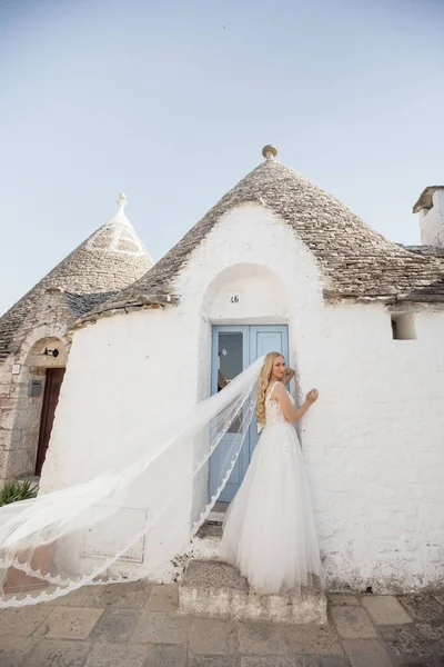 Beyaz gelinlik ve duvak takmış, gülümseyen ve İtalya 'da taş şehir evinin yanında duran genç sarışın gelin portresi. Güzel ve romantik bir düğün, sokakta yürümek..