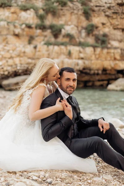 白い服を着た若いブロンドの花嫁とスーツを着たブルネットの新郎の肖像画は イタリア側のビューで岩の多い海の海岸に座って 背景を海景 美しくロマンチックな結婚式幸せなカップル — ストック写真