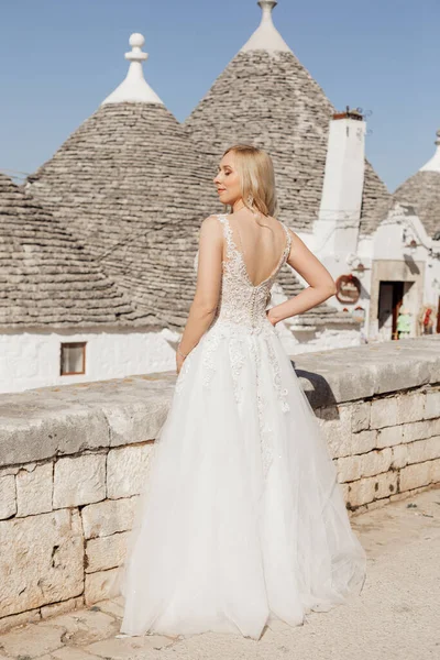 Elegant pretty blond woman, bride in wedding dress standing alone, posing against historical center of Italy with house roofs and sky. Stay with close eyes, female in wedding dress, joy sun. Rearview