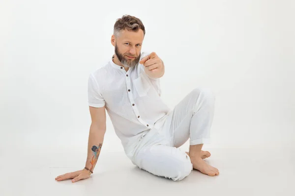 Bearded businessman with tattoo sitting on the floor in white outfit on white isolated background. Point at camera by forefinger, successful gesturing. Your turn, next. Are you ready, hey. Copy space