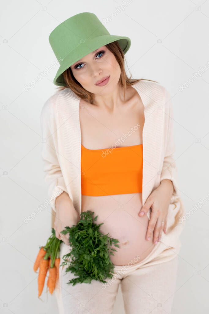 Woman with bare belly in late pregnancy holding carrot bunch in casual clothes with panama on head, white background. Portrait of young pregnant lady expecting baby closeup. Happy pregnancy concept.