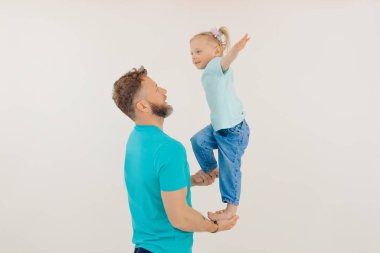 Mavi tişört ve kot pantolon giyen aile manzarası. Kısa saçlı, erkek eli üzerinde duran, elini kaldıran gülümseyen küçük kız. Baba kız bacağı tutuyor, eğleniyor, oynuyor. - Aile. Stüdyo.