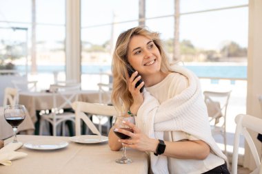 Beyaz tişört giyen, süveter giyen, beyaz sandalyede oturan, barajdaki restoranda şarap bardağını tutan, akıllı telefondan konuşan mutlu genç bir kadının portresi..