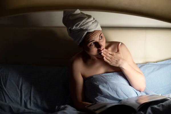 Portrait of young frightened afraid scared woman with white towel wrapped around head lying on bed covered with blue blanket, looking aside, covering mouth with hand, reading magazine. Overemotive.