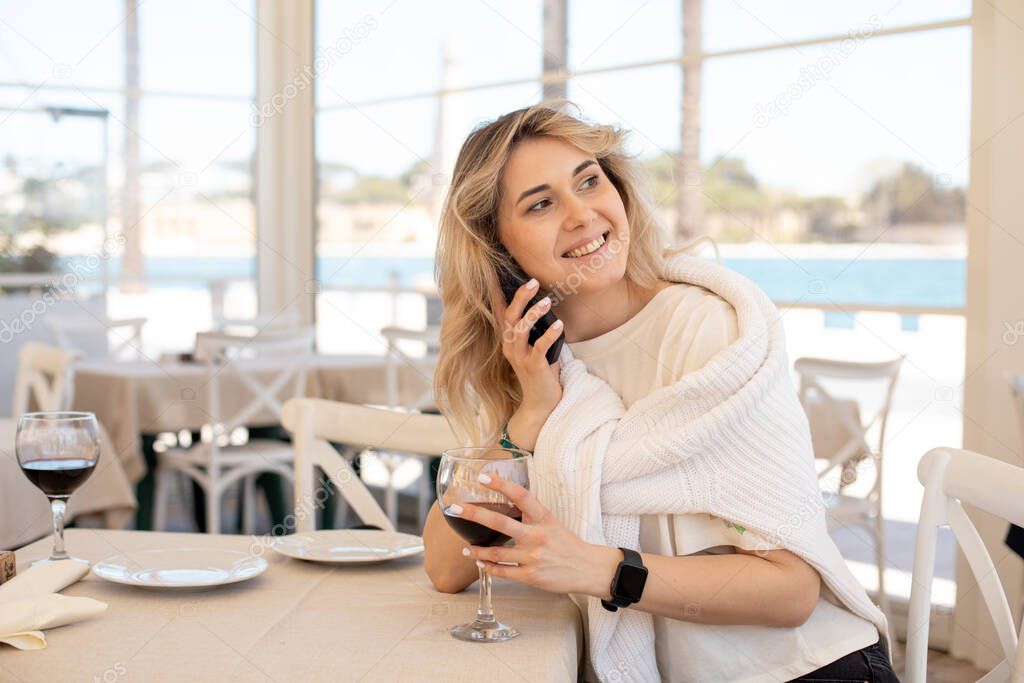 Portrait of young smiling happy woman wearing white T-shirt, sweater, sitting at table on white chair, holding glass of wine in restaurant cafe on embankment, talking on smartphone, looking aside.