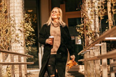 Zarif iş kadını şişirilmiş süveter ve siyah ceket kahve molasında elinde eklenebilir bir fincan kahveyle yüzünde sonbahar güneşinin sıcaklığıyla yürüyüşe çıkıyor. Yaşam biçimi.