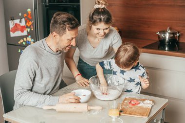 Sevimli genç anne zevkle bakıyor. Erkekleri kurabiye pişirmek için hamur yoğuruyor. Baba ve oğul takım olarak çalışırlar, anne onlara yemek yapmayı öğretir. Ev mutfağı. Takım çalışması. Anne, baba, oğul