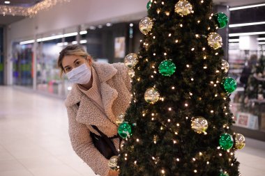 Güzel bir kadın alışveriş merkezinde süslü Noel ağacının arkasından bakarken eğleniyor. Tıbbi maskeli kadın. Noel 'de alışveriş merkezi. Yeni normal konsept. Tatillerde. Yeni yıl havası..