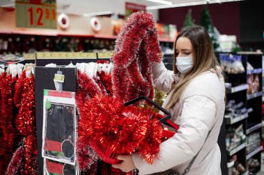 Beyaz kışlık ceketli ve alışveriş merkezinde koruyucu maskeli güzel bir kadın Noel için evi dekore etmek için kırmızı çelenkler alıyor. - Evet. Noel alışverişi. Kişisel koruma ekipmanları.