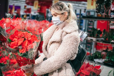 Açık renk ceketli ve maskeli güzel bir kadın süpermarketin bitki reyonunda çiçek açan kırmızı noktalıyı seçiyor. Kişisel koruyucu ekipman. Noel yıldızı. Noel ve yeni yıl..