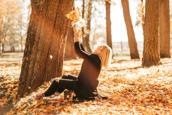 Happy woman throws fallen yellow leaves into air on sunny autumn day sitting on old suitcase in park. Fun while hiking in autumn season. Lifestyle. Colors of autumn. Leaf fall.