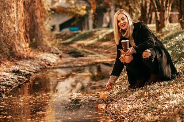Stylish blonde crouching near stream pulls her hand to water in autumn city park. She is illuminated by golden autumn light and holds cup with hot drink in her hands. Fabulous autumn landscape.