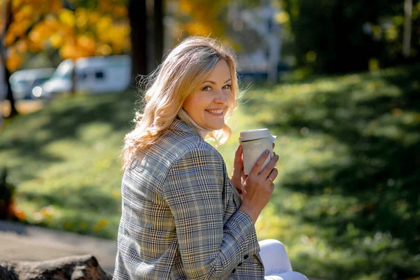 Beautiful blonde woman in light tweed jacket smiles looking over her shoulder at camera. Autumn day in nature. Happiness and peace. Life in moment. Walking in fresh air. Coffee break.