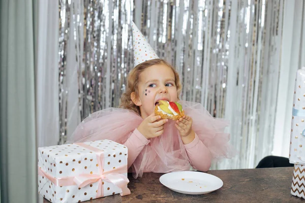 Charming 4-5-year-old baby in party hat and chiffon pink cape with decorative sequins on her face celebrates her birthday. She is sitting attable and enjoying birthday cake, next to gift box.