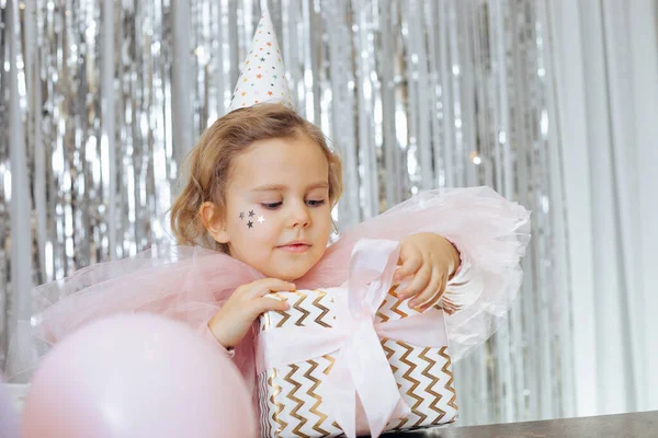 Birthday. Gifts for children. Charming girl in party hat and pink airy outfit is completely passionate about unpacking beautifully designed gift. Gift delivery. Happy childhood. Pink and silver.
