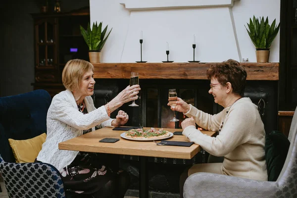 Two senior women friends having dinner at restaurant, attractive lady spending time together indoors. old friends drinking wine in modern apartment. Celebration.