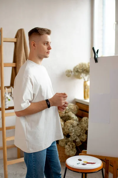 Blonde casual clothed young man standing and drawing. Artist painting on canvas, doing arts. Male holding brush and pallette with colors. Painter standing in light white studio, doing his hobby.