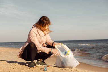 Kadın eylemci ve asistan, kumlu plajı su altındaki çöplerden temizliyor. Deniz ya da okyanus, plastik atıklarla. Poşetlemek için şişe toplayın. Ekolojik kirlilik. Sürdürülebilir kalkınma, küresel değişim. Rüzgarlı