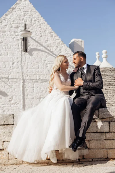 恋にロマンチックな結婚式のカップルの垂直肖像画 ブロンドの女性と黒髪の男性の結婚式後 服を着て お互いを受け入れる見て 一緒に古代イタリアの町に対して石の岩の壁に座って — ストック写真