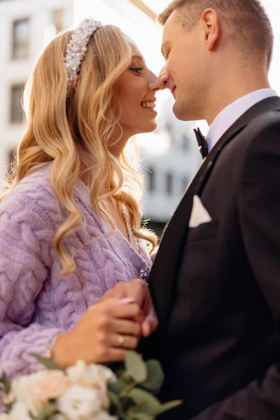 Beautiful young wedding couple in elegant outfits walk around city, smile and kiss on street background. Portrait side view closeup of happy bride and groom in love. Wedding day, family holiday.