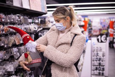 Kış kıyafetleri ve maskeli güzel bir kadın yeni yıl satış departmanındaki süpermarkette sevimli bir Noel oyuncağı tutuyor. Yeni yıl. Salgın hastalıklar. Yeni yıl alışverişi. Noel ağacının altında oyuncaklar..
