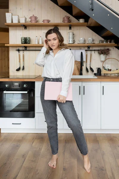 Portrait of serious dark-haired woman in white shirt and jeans, standing barefoot on wooden floor in front of her kitchen and holding laptop. Work at home online. Kitchen designer. Lifestyle.