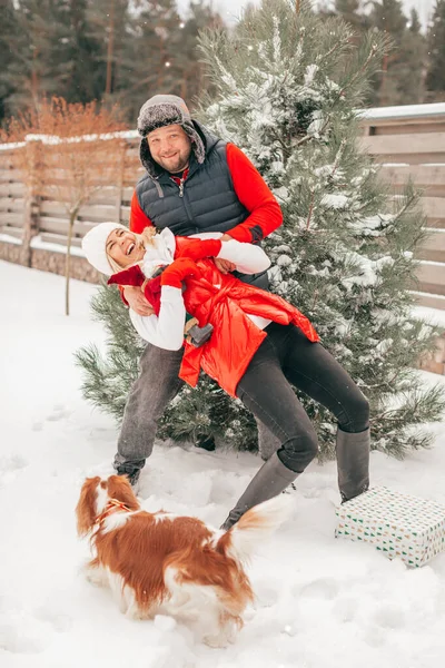 man and woman playing with snow in snow-covered front or back yard, people have fun on winter day, dog looking on them. Happy couple