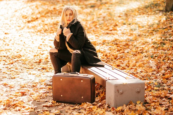 秋は変化の時です 魅力的で 黒いカシミアコートとジーンズの女性的なブロンドは都市公園の庭のベンチに座っており 古いスーツケースは彼女の隣に立っています 今の生活 ライフスタイル — ストック写真