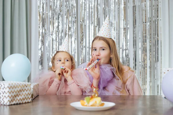 Two sisters of 5 and 11 years old in party hats and elegant dresses are sitting at table and blowing holiday horns, with cake in front of them. Birthday. Family values. Happy childhood.