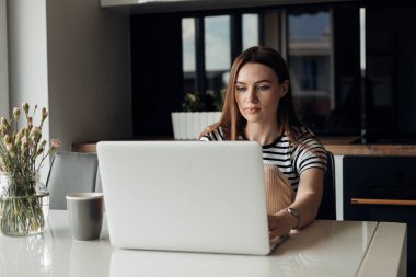 Masada oturan, çay fincanının yanında beyaz dizüstü bilgisayarda, çiçek karanfilli vazo yazan genç, konsantre olmuş güzel bir kadının portresi. Uzak ofis, serbest meslek, internet, uzaklık öğrenimi.