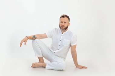 Sakallı, çıplak ayaklı, düşünceli, dövmeli, beyaz elbiseli, izole edilmiş arka planda oturan, kameraya bakan ve düşünen bir adam. Küçük işletme, yoga ve spor salonu koçluğu.