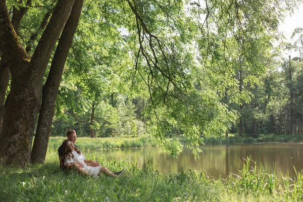 Young couple falling in love sitting on grass under tree near lake in park, looking on each other. Young man and woman enjoying summer day. Happy people in love, honey moon. Lifestyle.
