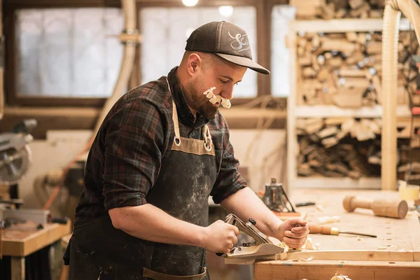 Concentrated, playful man, carpenter in cap and protective apron with funny shaving chip mustache learning to work with jack plane. Making, framing wooden natural furniture on bench table in workshop