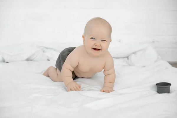 Cheerful little baby crawl on big bed, have fun and laugh on white background. Portrait of beautiful naked infant child, free copy space. Concept of happy childhood and caring for children.