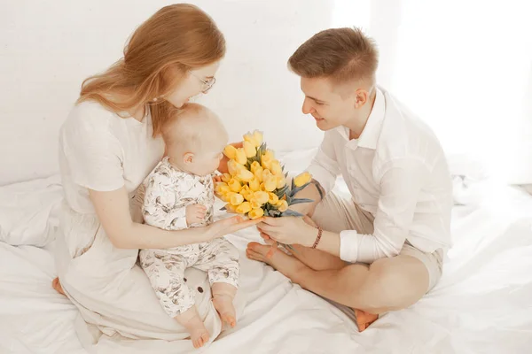 若い母親と父親はベッドの上に小さな赤ちゃんと一緒に座って 男は白い背景に黄色のチューリップの花束を与える 親と幼児のかわいい関係 幸せな家族と親の愛情 — ストック写真