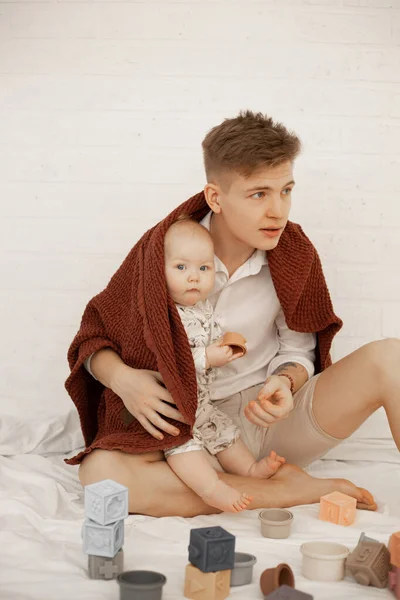 stock image Young father embrace baby, sit under brown towel on bed and play toy cubes on white background. Tender relationship between man and infant child, free copy space. Concept of parental affection.