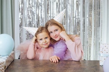 Doğum günlerini kutlayan iki sevimli kız kardeşin portresi. Parti şapkalı ve şenlikli elbiseli kızlar yanakları elleriyle desteklenmiş şekilde aynı şekilde otururlar. Mutlu bir çocukluk. Aile değerleri. 