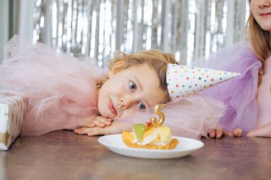 Sevimli beş yaşındaki kız başını avuçlarına koydu ve doğum günü pastasına hayranlıkla baktı. Neşeli çocuklar duyguları etkiler. Küçük kız ve doğum günü pastası. Aile tatilleri.