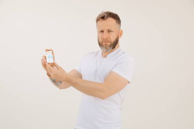 Beyaz tişört giyen orta yaşlı sakallı bir adamın portresi. Beyaz arka planda etki hapı içeren plastik bir şişe taşıyor. İktidarsızlık, erkek cinsel sorunları için ilaç. Stüdyo.