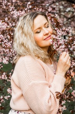 Uzun, açık renk saçlı, pembe hırka giyen, çiçek açan kiraz ağacının yanında duran, parkta dal tutan, beyaz çiçeği koklayan güzel bir kadının yan görüntüsü. Bahar. 