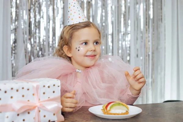 Smiling baby 4-5 years old in party hat and chiffon pink cape with decorative sequins on her face is sitting at table on which there is birthday cake and gift box. Birthday. Happy childhood.