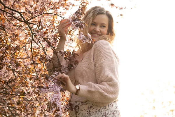 穿着粉色羊毛衫的年轻美丽的金发女子的画像 夏天印花的衣服站在开着花的樱桃树旁 在公园里拿着枝白花的枝条 — 图库照片