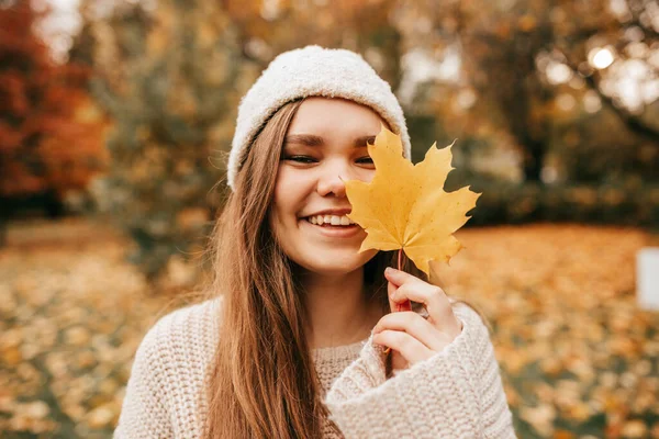 Güz ormanında örülü şapka ve süveter giyen, elinde akçaağaç yaprağı tutan, düşen parkta yürüyen, kameraya bakan, mutlu insanlar.