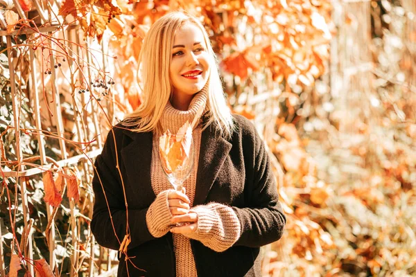 黒コートとベージュのセーターの美しいブロンド35歳は彼女の手に秋のカクテルとガラスを保持し 都市公園で晴れた日を楽しんでいます 秋の色 休息と喜び 葉の秋 — ストック写真